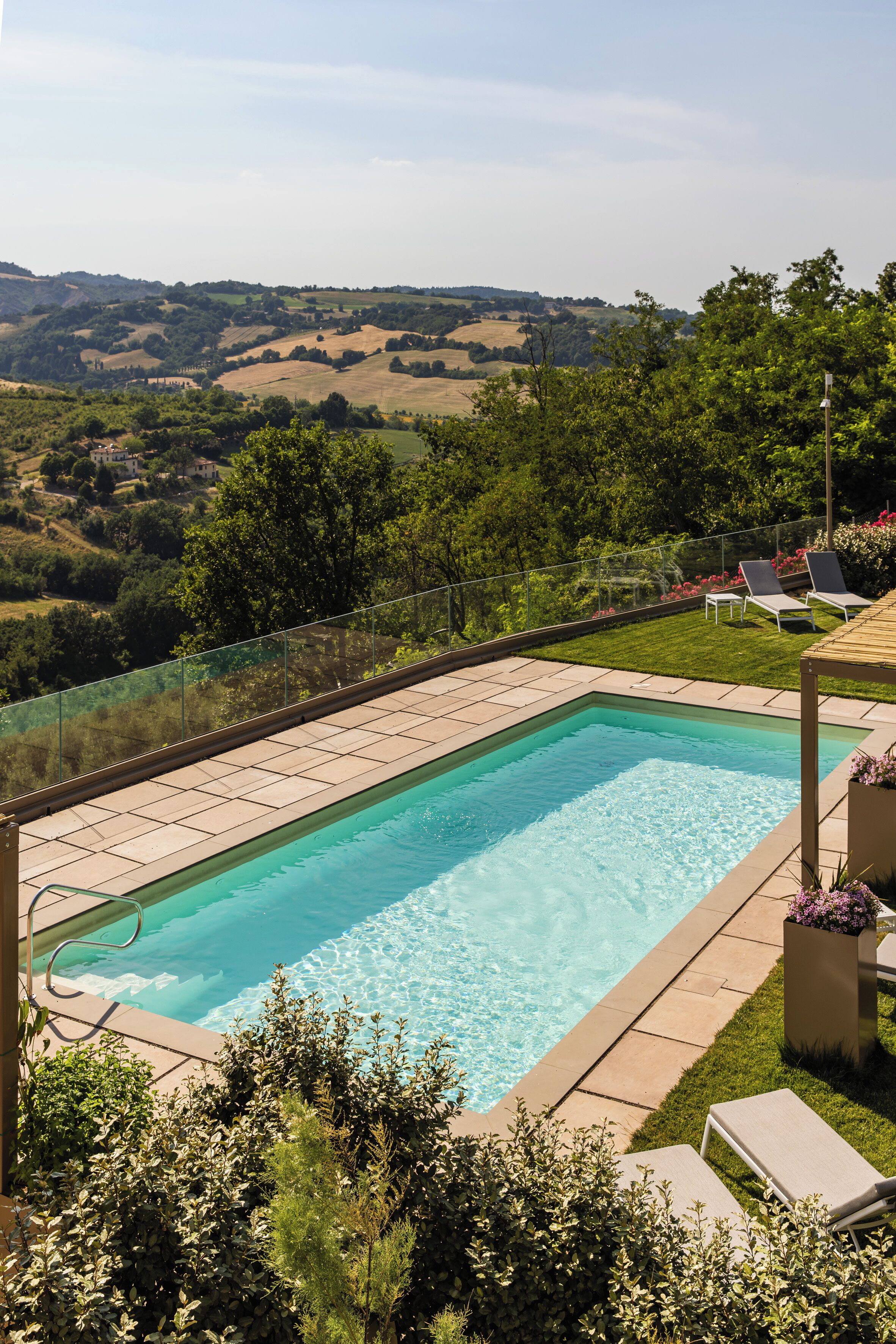 A rectangular swimming pool with clear blue water, surrounded by a paved deck and a glass balustrade.