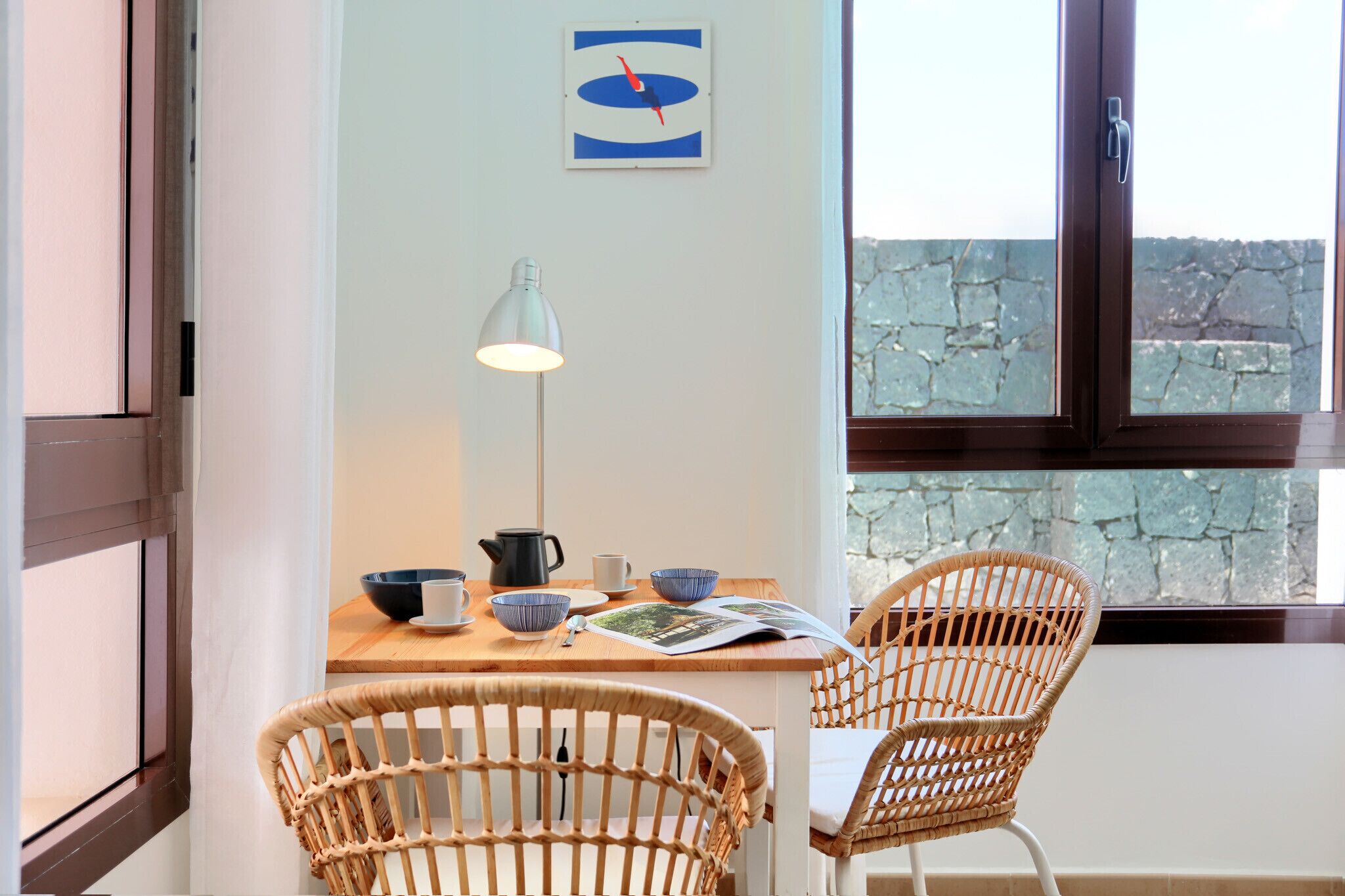 Un coin repas avec des chaises en osier, une table sur laquelle sont posés des bols, une bouilloire et une lampe.