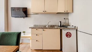 En kompakt tekøkken med et TV ophængt over bordpladen, et lille køleskab med logo, og et spiseområde med en grøn stol.