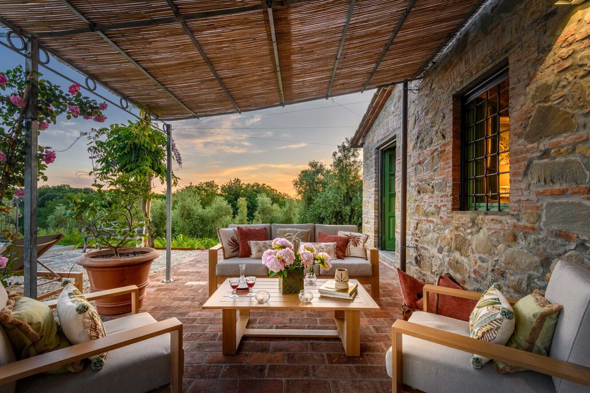 A covered outdoor seating area with a stone building, a table with drinks, and potted plants.