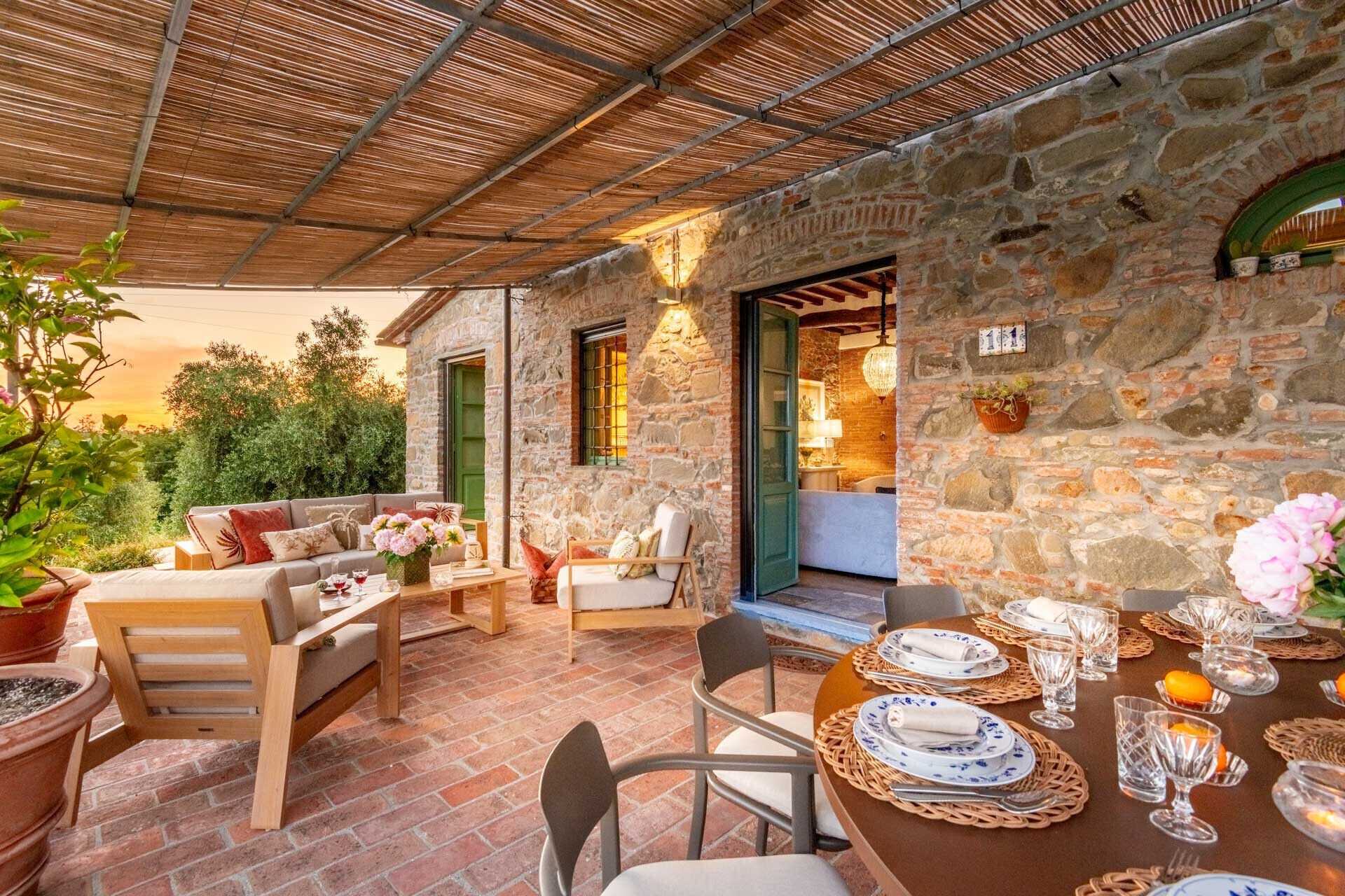 Un área de comedor al aire libre con paredes de piedra, una mesa dispuesta para una comida, rodeada de asientos y plantas en macetas.