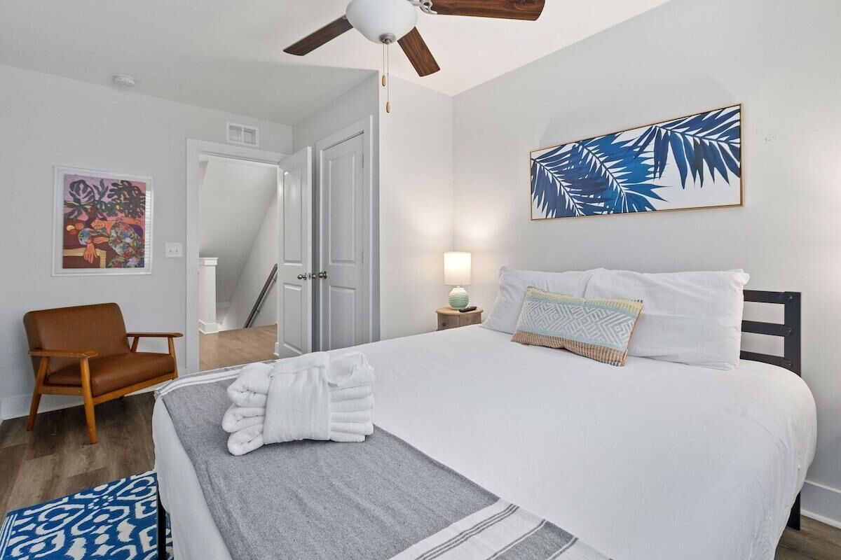 A bedroom with a bed, a chair, a ceiling fan, a wall art piece, and a door.