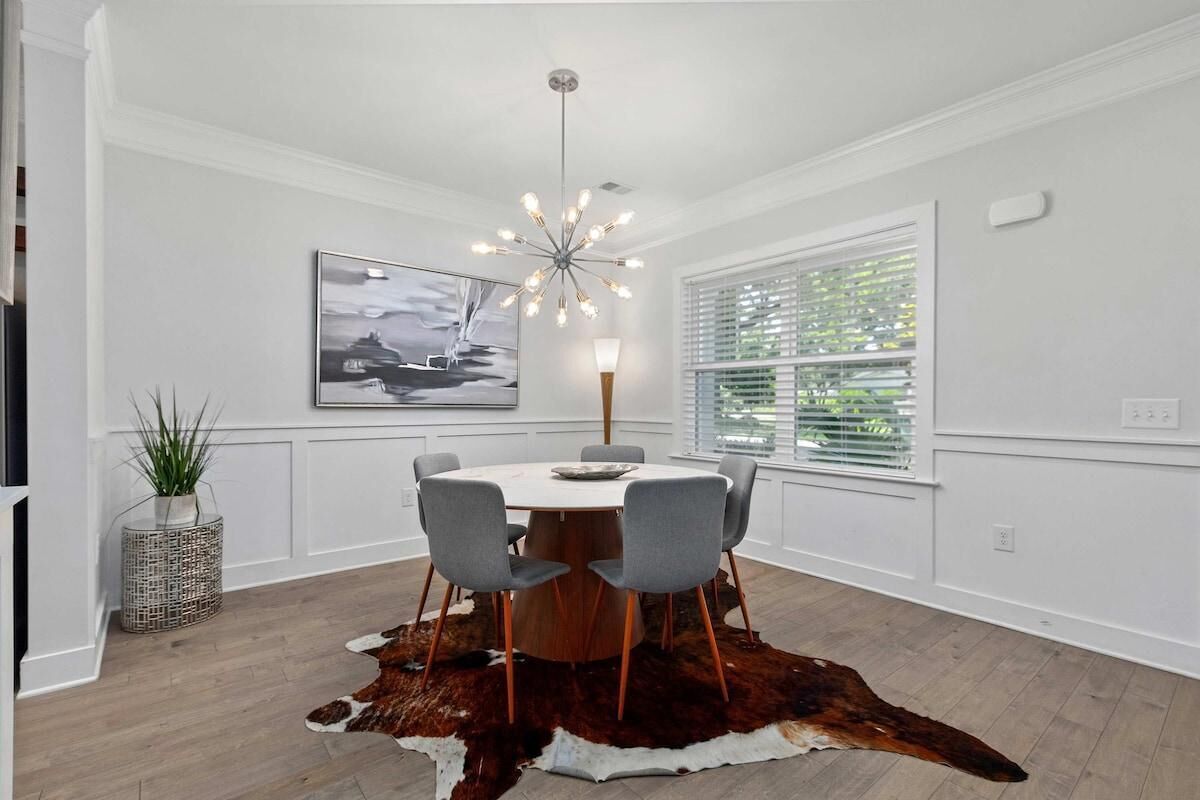 Een eetruimte met een ronde tafel, grijze stoelen, een koeienhuidtapijt, een moderne kroonluchter, een aan de muur bevestigde televisie en een raam met blinds.
