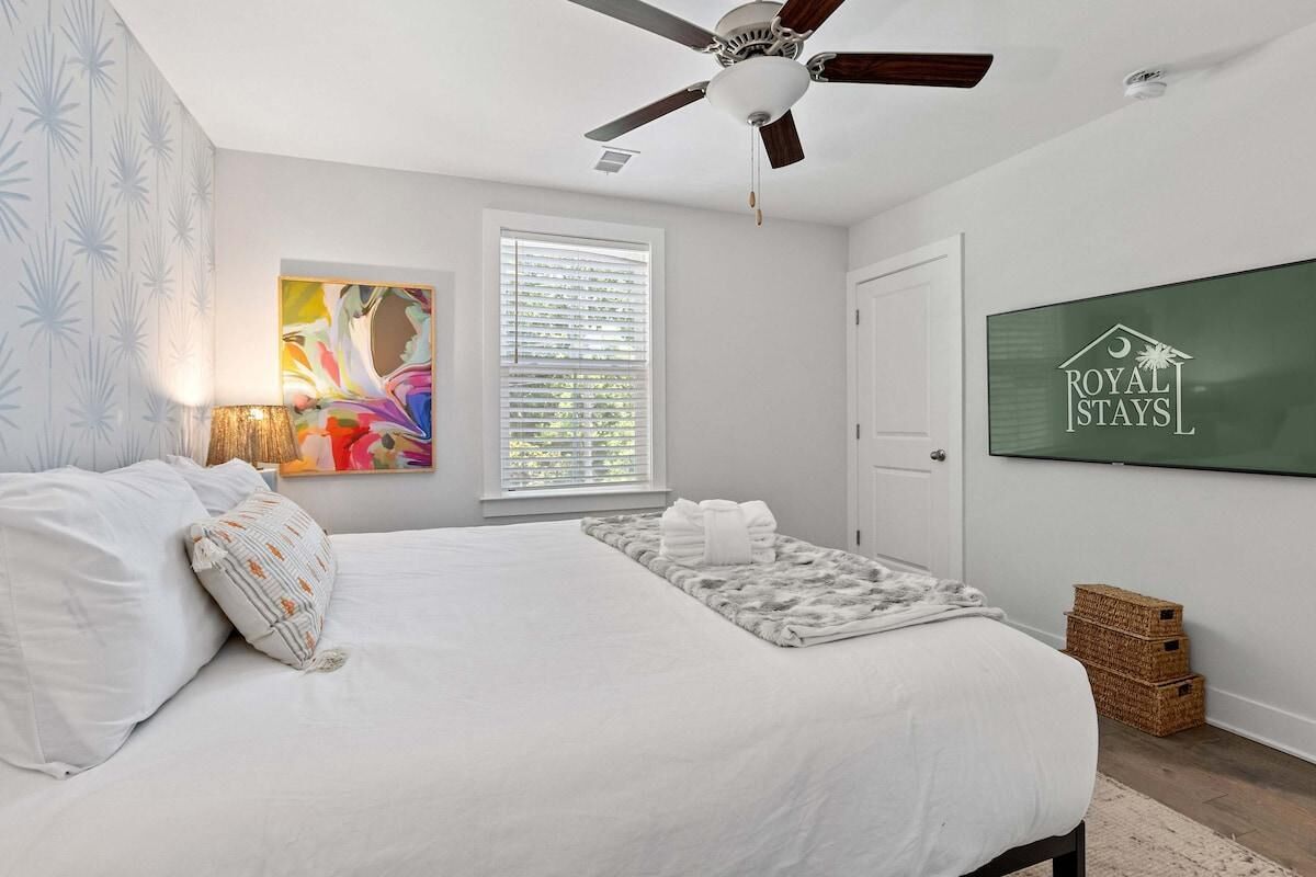 A bedroom with a bed, a ceiling fan, a window with blinds, a wall-mounted artwork, and a sign for Royal Stays.