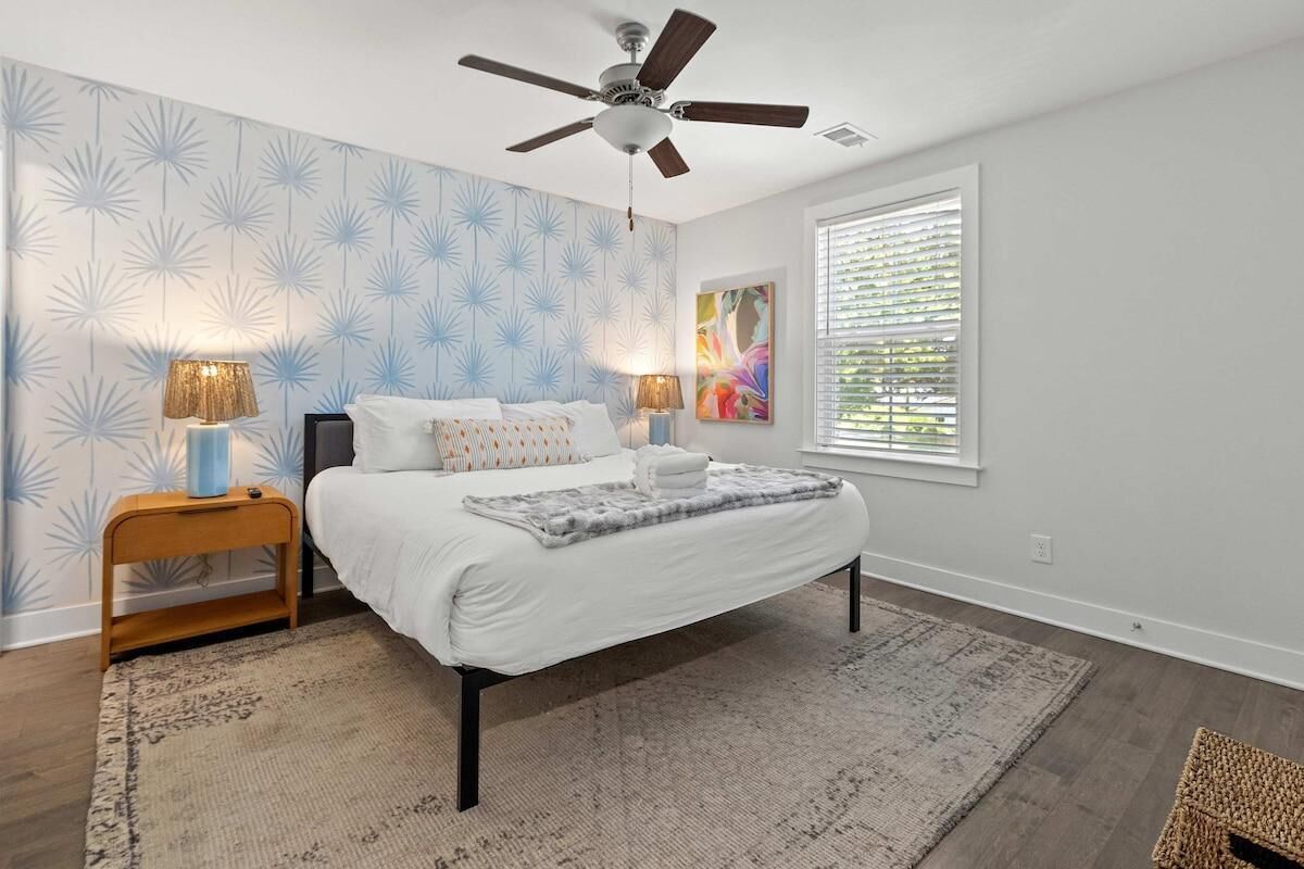A bedroom with a bed, two bedside lamps, a ceiling fan, a window with blinds, and a patterned rug.