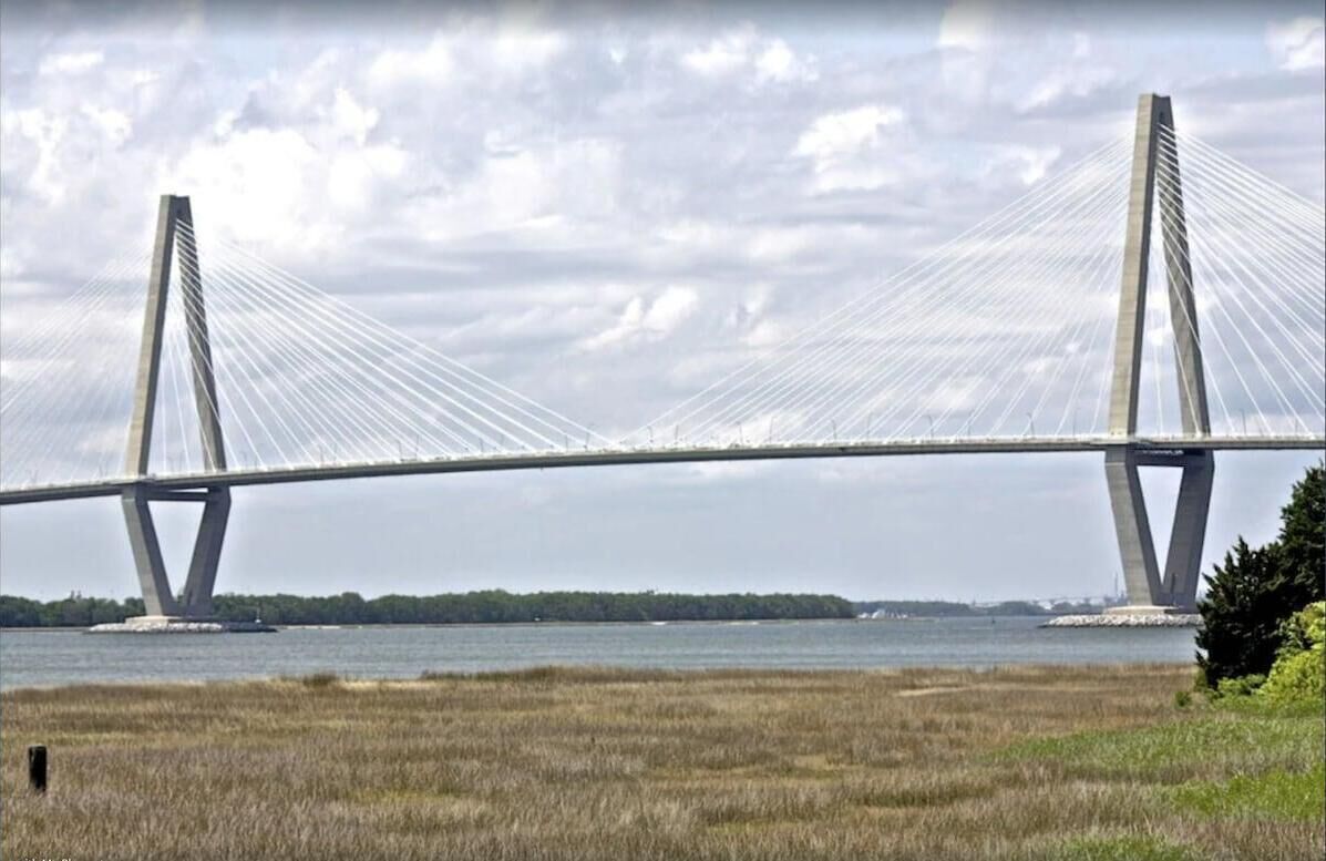 A modern bridge with a cable-stayed design spanning over a body of water, with a grassy foreground and a cloudy sky in the background.