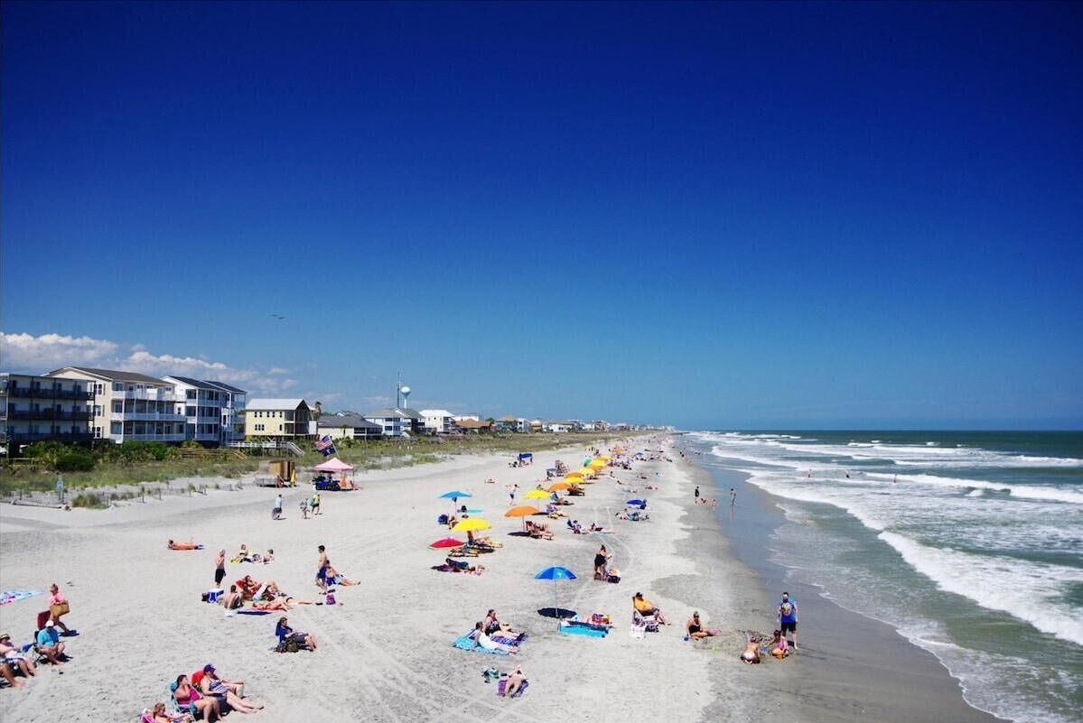 A beach with people sunbathing and swimming, umbrellas, and buildings in the background.