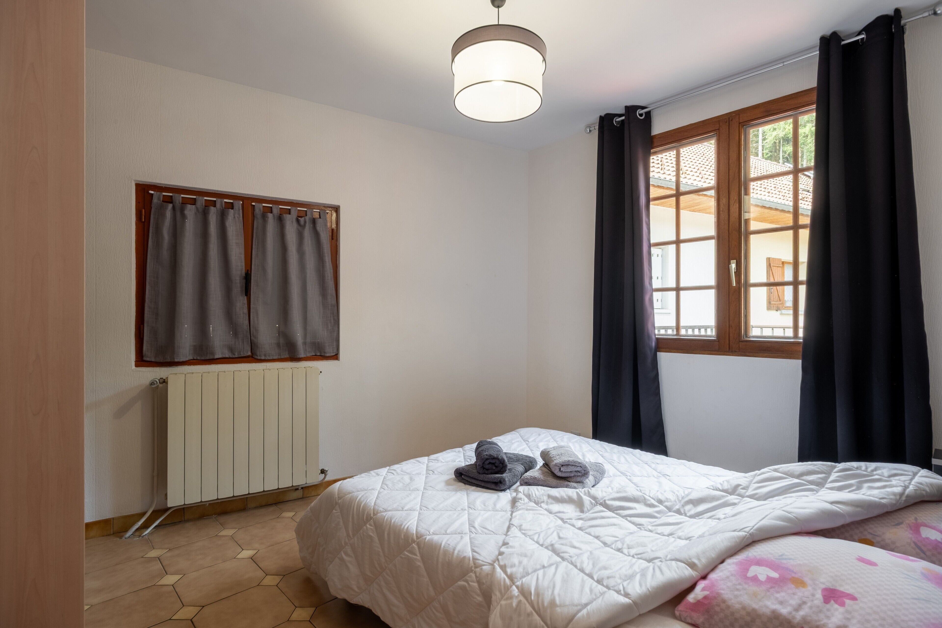 A hotel room with a bed, a radiator, a window with curtains, and a ceiling light.