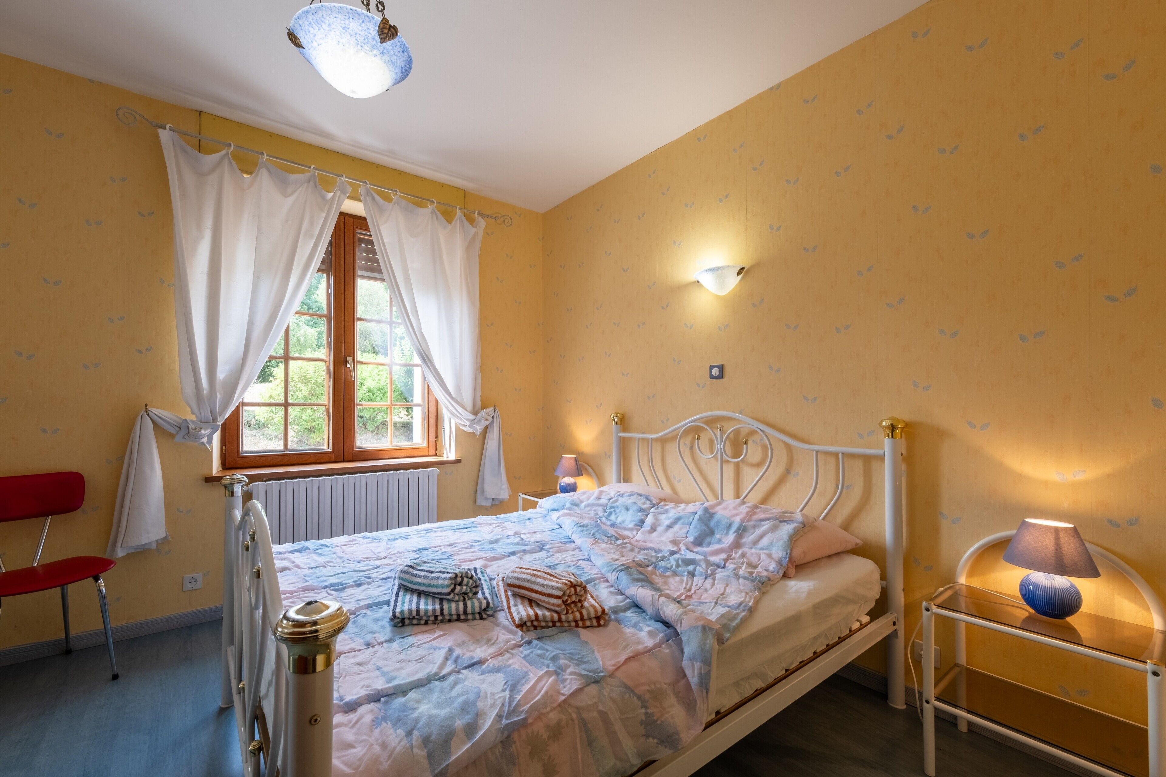 Une chambre à coucher avec un lit, une fenêtre avec des rideaux, un radiateur, une table de chevet avec une lampe et une chaise.