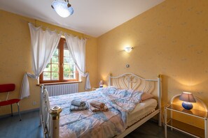 A bedroom with a bed, a window with curtains, a radiator, a bedside table with a lamp, and a chair.