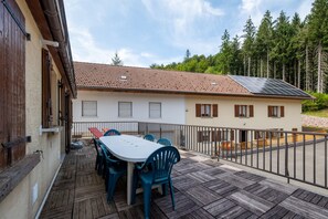 A patio with a table and chairs, a building with a tiled roof, and a forest in the background.