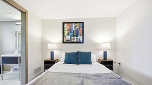 A hotel room with a bed, two bedside tables, a nightstand, a bathroom with a mirror, and a cityscape painting on the wall.