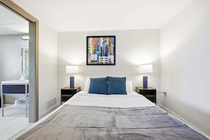 A hotel room with a bed, two bedside tables, a nightstand, a bathroom with a mirror, and a cityscape painting on the wall. - 2 BR Iconic Detroit High-Rise Loft (Detroit)