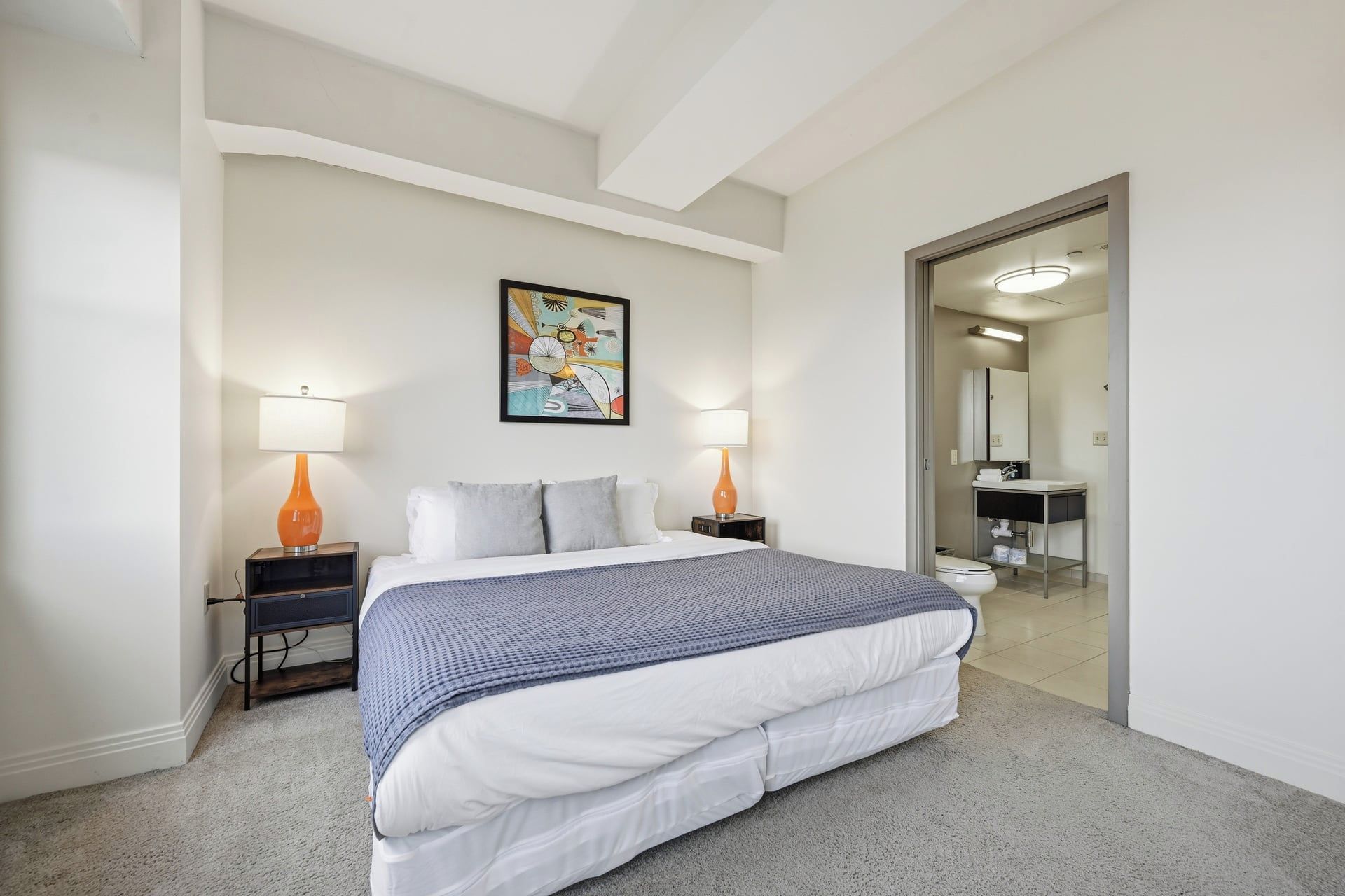 A hotel room with a large bed, two bedside tables with lamps, a painting on the wall, and a bathroom visible through an open door.