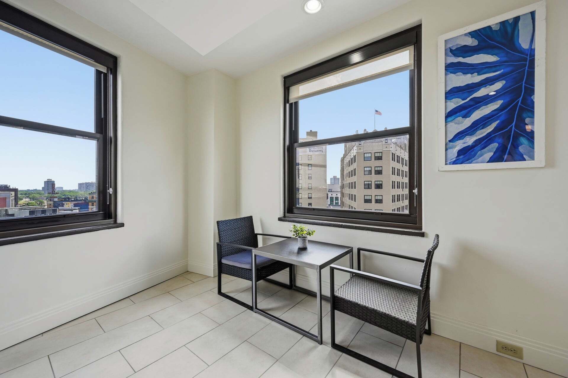 A room with a table and two chairs, a window with a view of buildings, and a blue abstract painting on the wall.