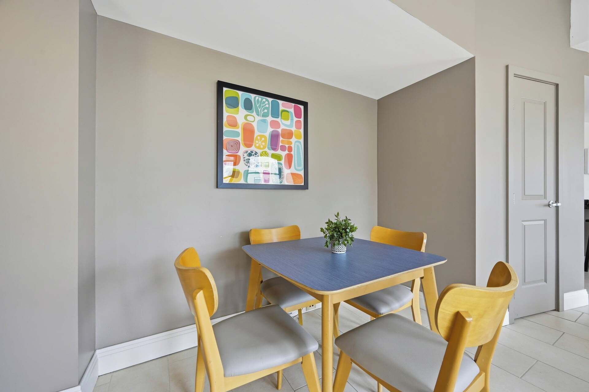 A dining area with a table and four chairs, a potted plant on the table, and a colorful abstract painting on the wall.