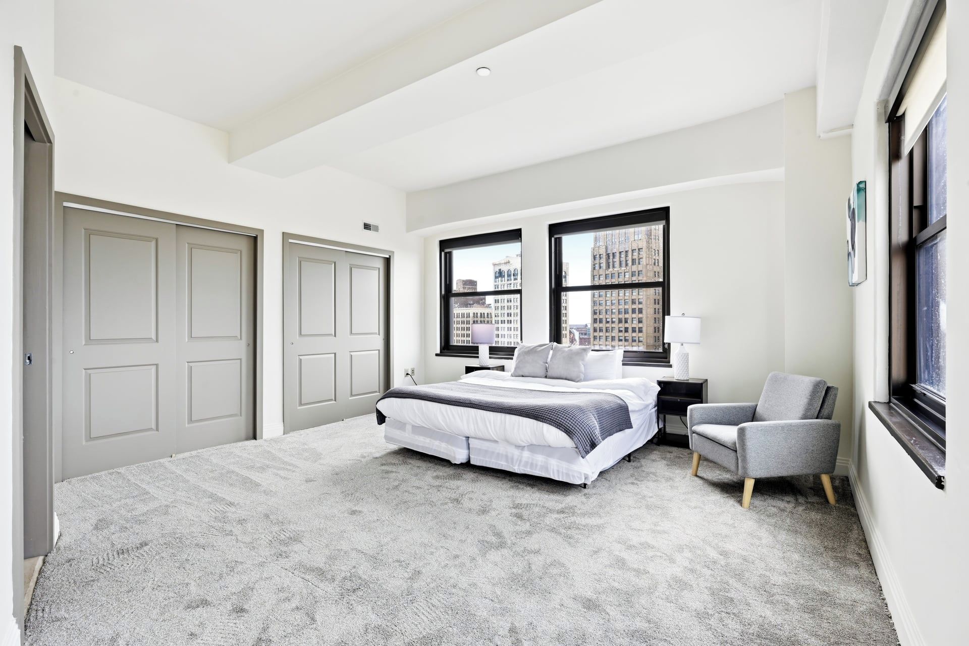 A hotel room with a large bed, two windows with city views, a chair, and a nightstand.