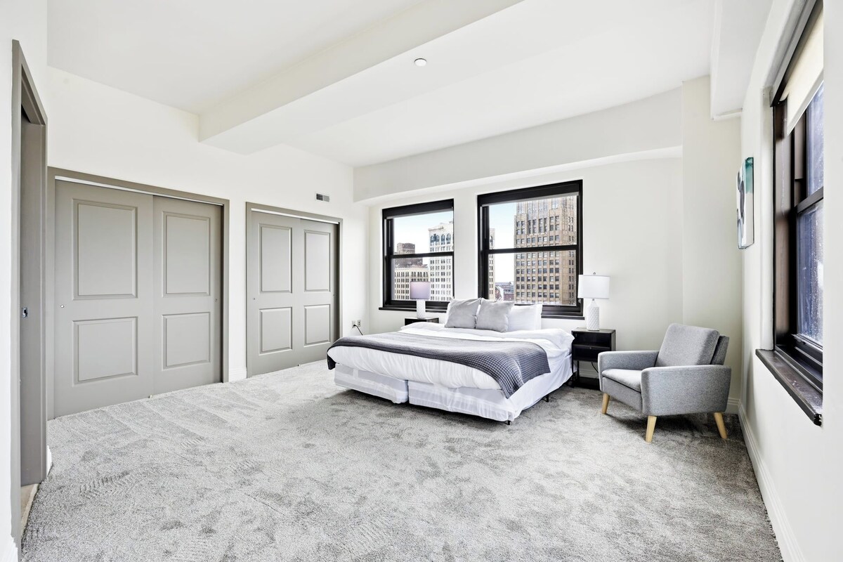 A hotel room with a large bed, two windows with city views, a chair, and a nightstand.