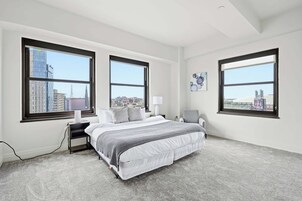 Quarto com uma cama grande, duas mesas de cabeceira, uma cadeira e três amplas janelas com vista para a cidade.