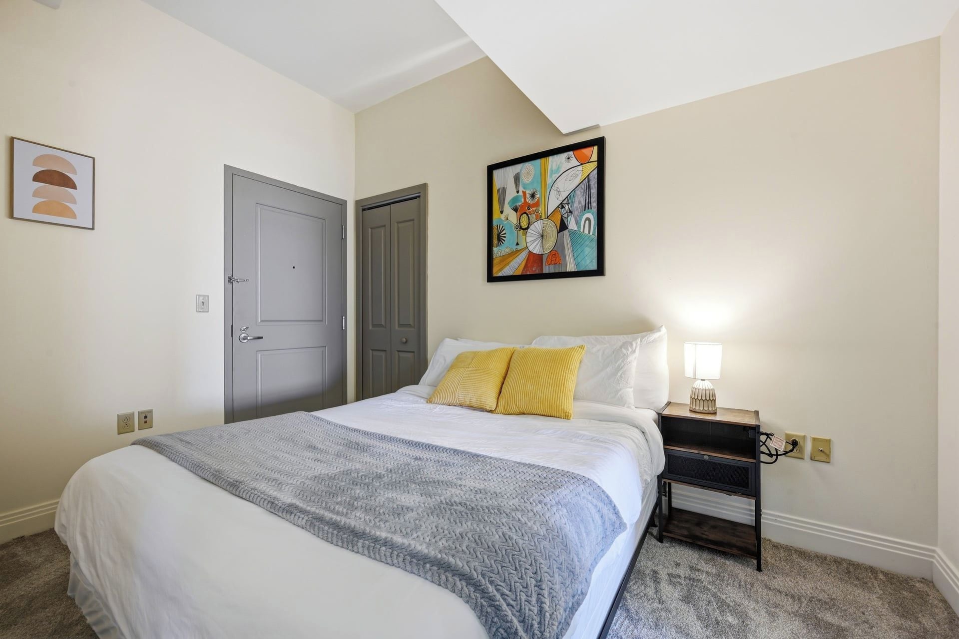 A bedroom with a bed, a nightstand, a lamp, a door, and a wall painting.