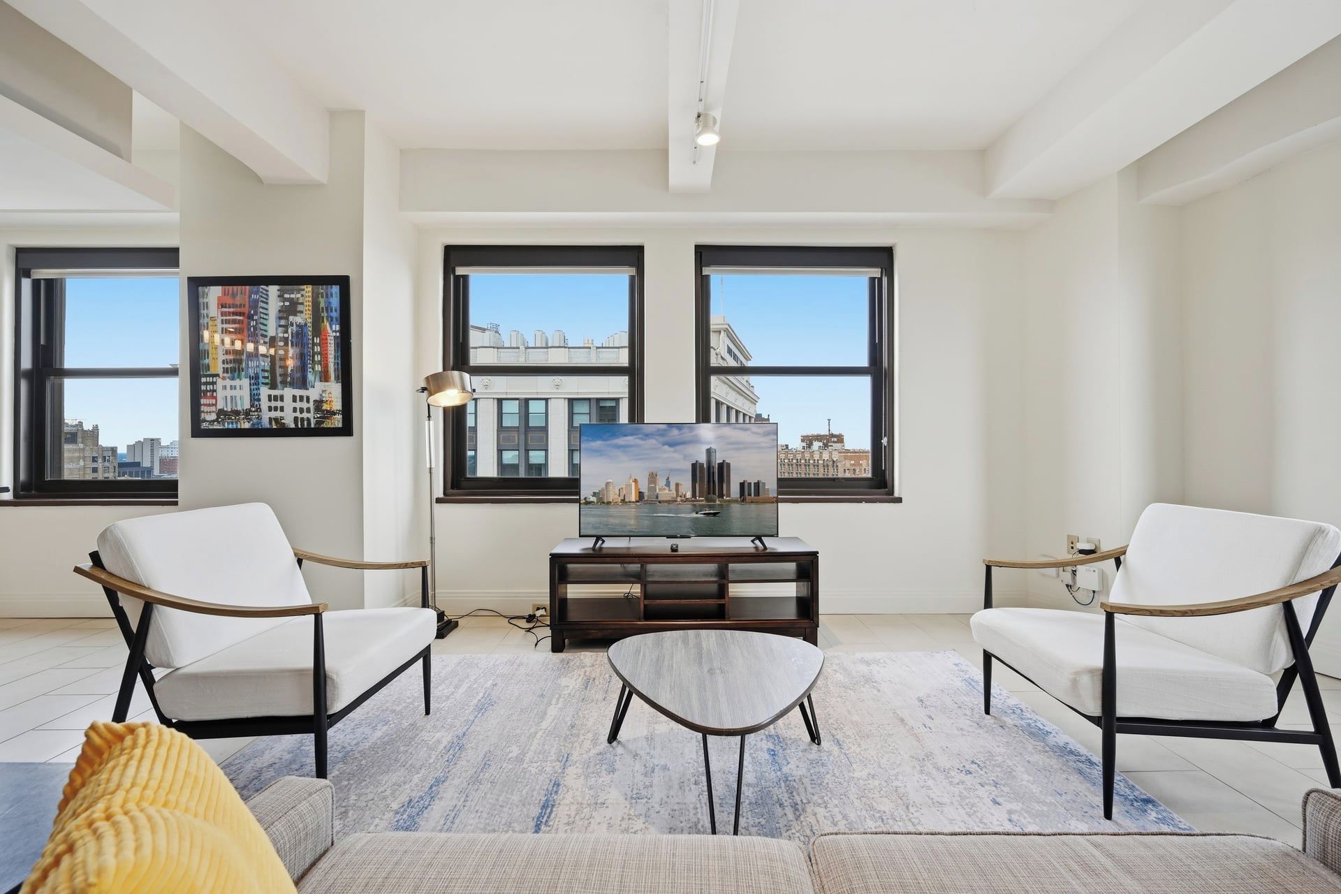 A modern living room with two white armchairs, a small coffee table, and a flat-screen TV on a wooden stand. Large windows offer views of a cityscape.