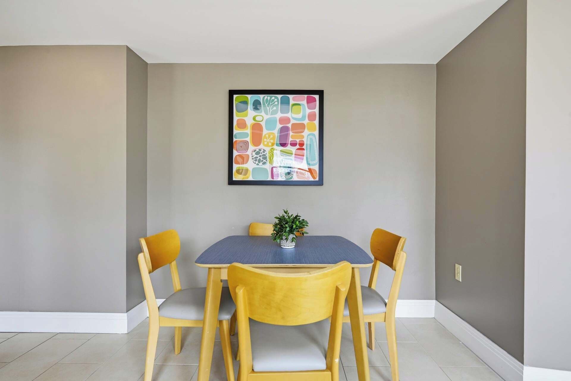 A dining area with a table and four chairs, a framed artwork on the wall, and a potted plant on the table.