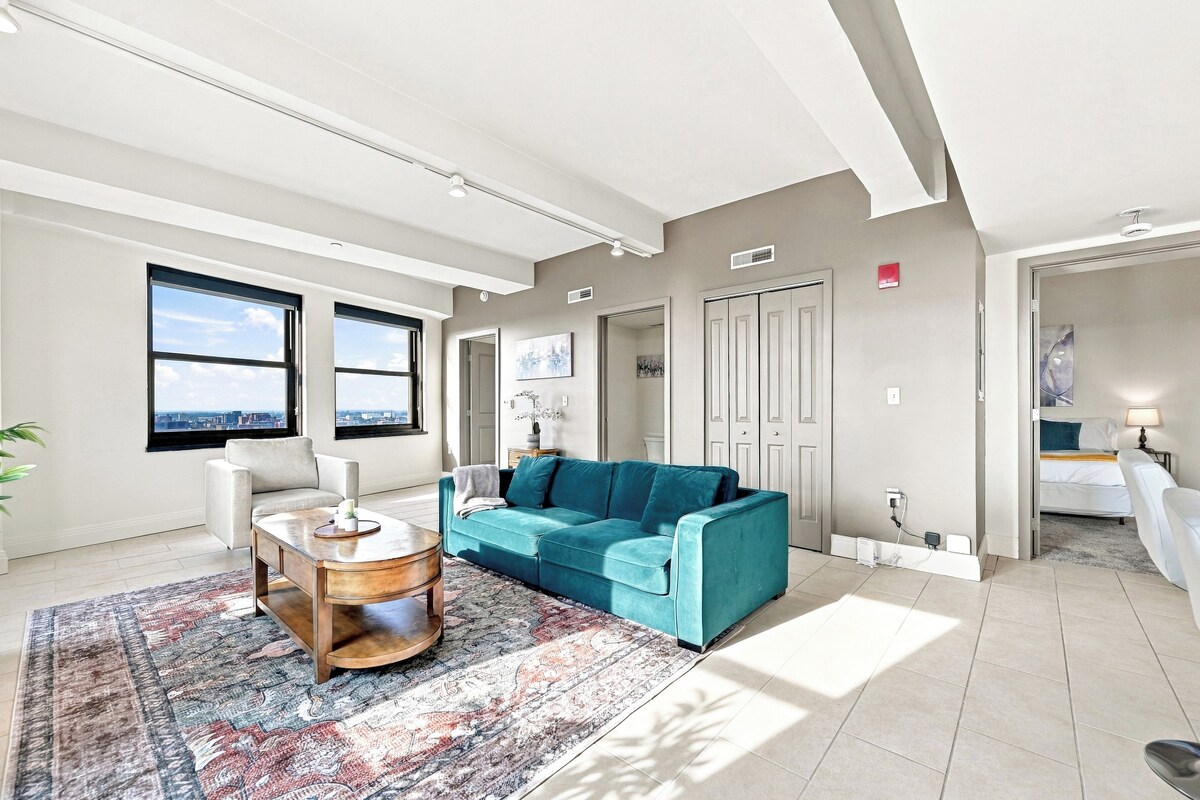 A spacious living room with a large window, a teal sofa, a coffee table, and a patterned rug.