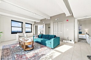 A spacious living room with a large window, a teal sofa, a coffee table, and a patterned rug. - 2 BR Iconic Detroit High-Rise Loft (Detroit)