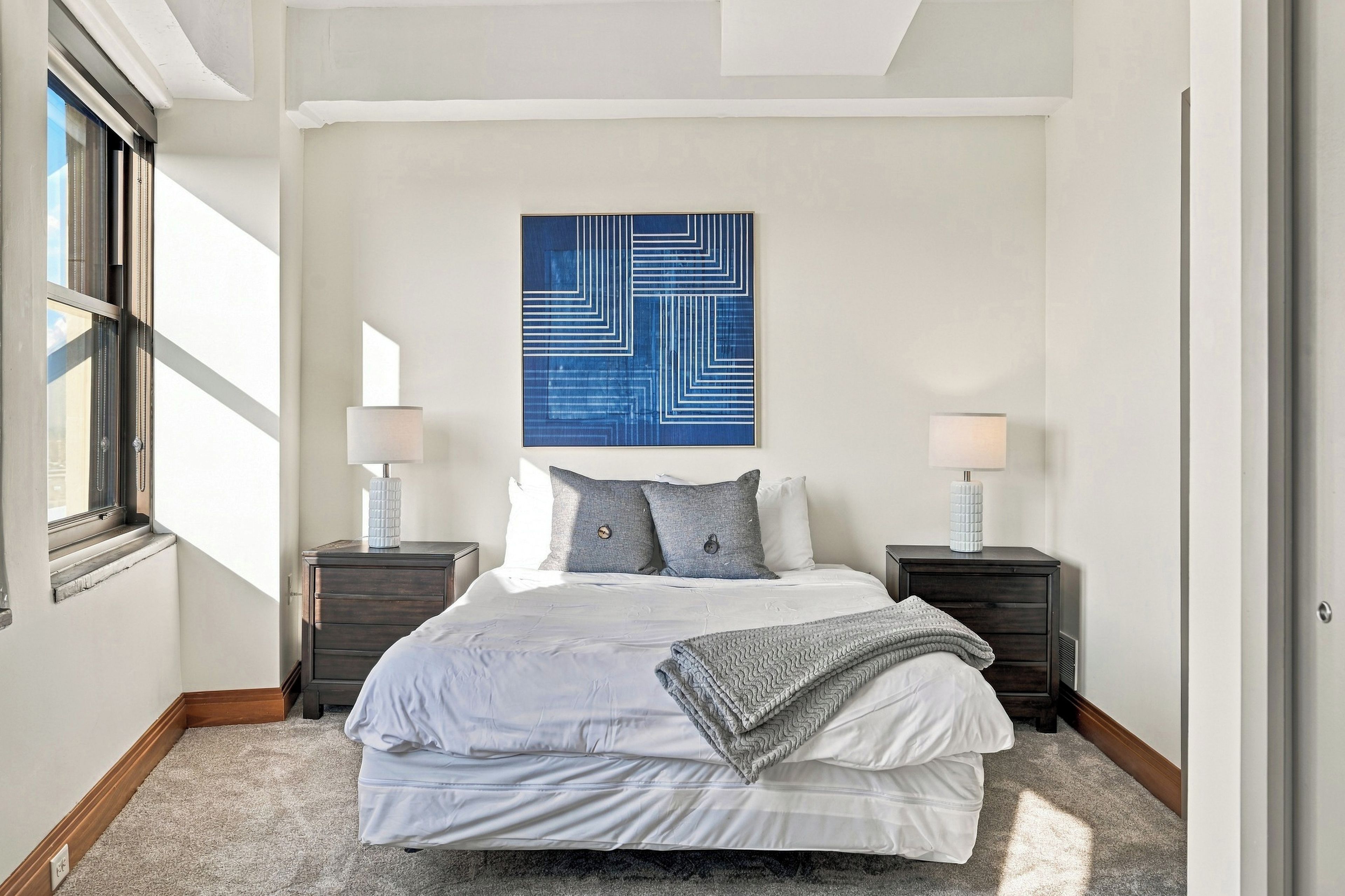 A bedroom with a bed, two bedside tables, a wall-mounted lamp, and a large abstract painting.