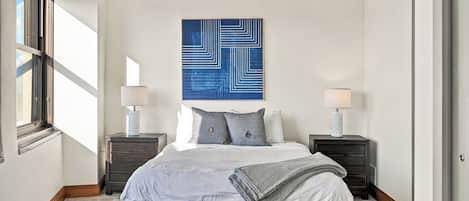 Une chambre à coucher comprenant un lit, deux tables de chevet, une lampe fixée au mur et un grand tableau abstrait.