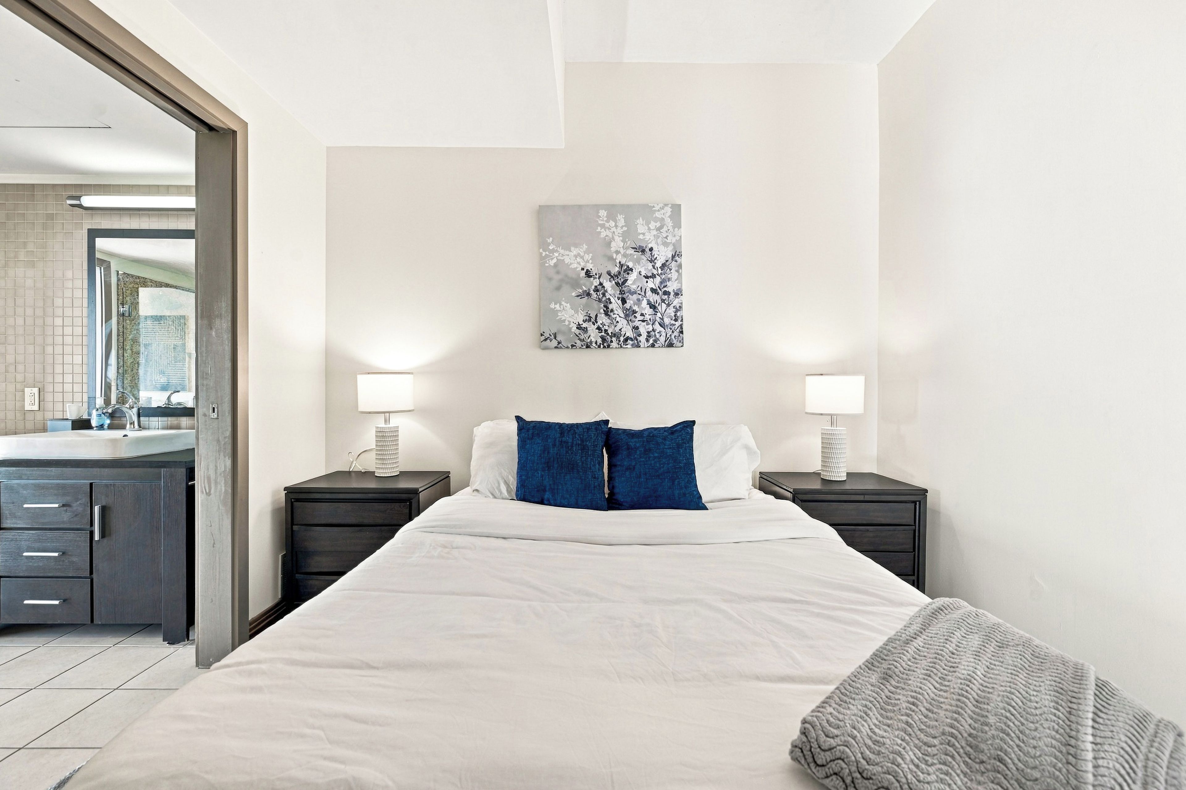 A hotel room with a large bed, two bedside tables with lamps, a wall-mounted artwork, and a view of the bathroom.