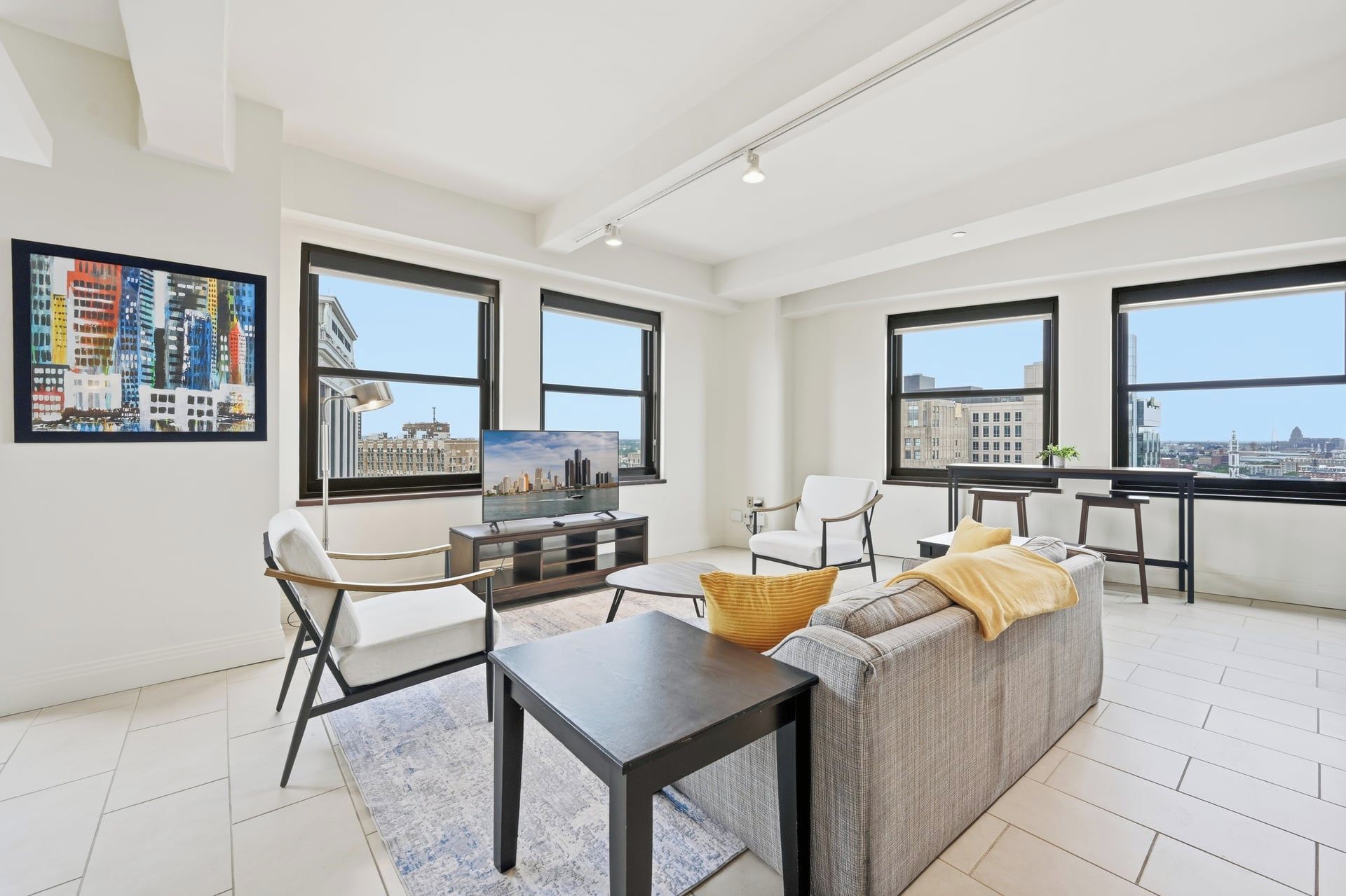 Un salon moderne avec un canapé, des fauteuils, une table basse et un téléviseur. De grandes fenêtres offrent une vue sur la ville.