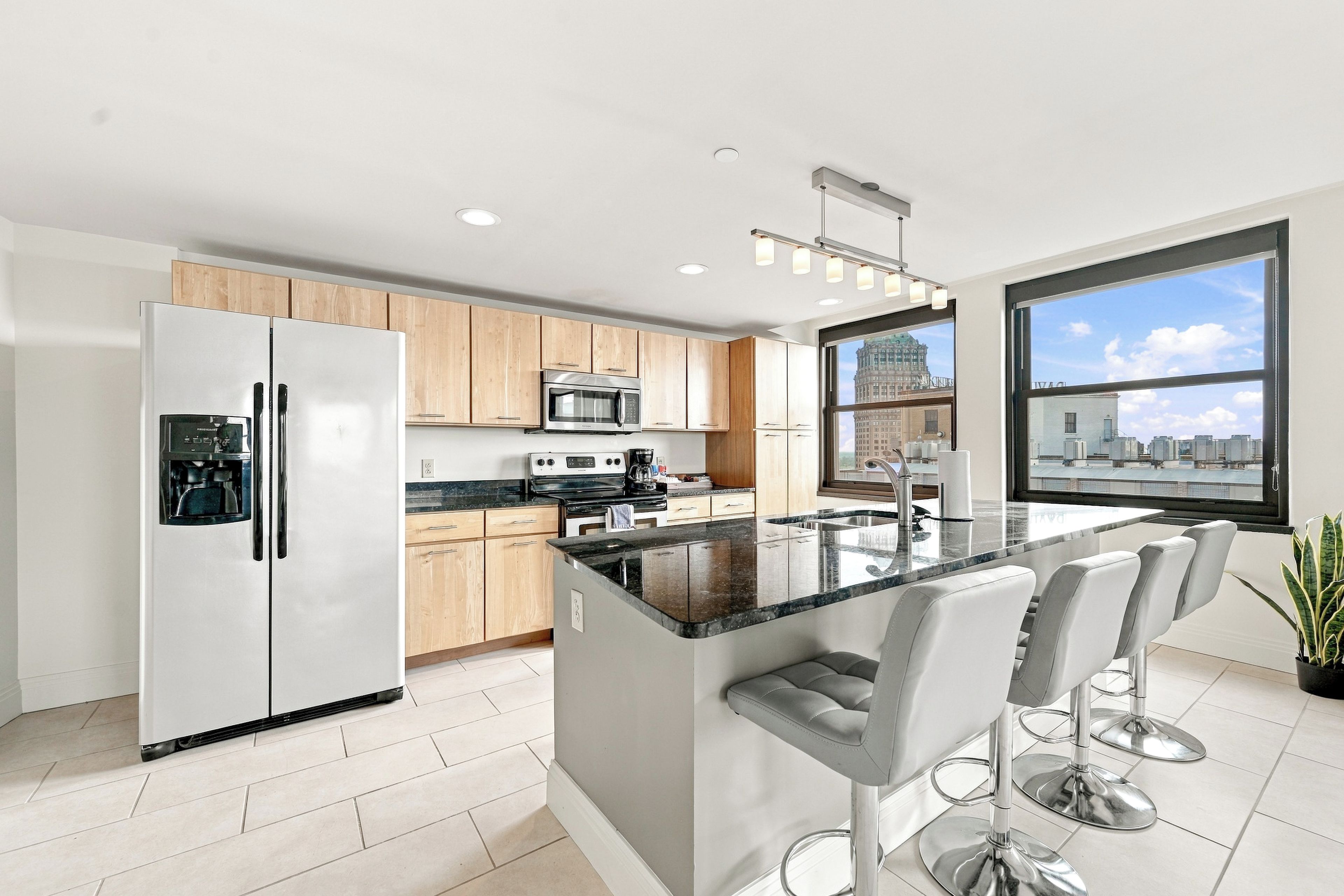 A modern kitchen with stainless steel appliances, a central island with bar stools, and a large window offering a city view.