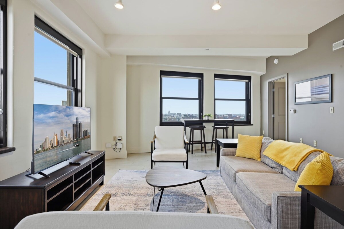 A modern living room with a sofa, a coffee table, and a TV. There are two windows with a city view.