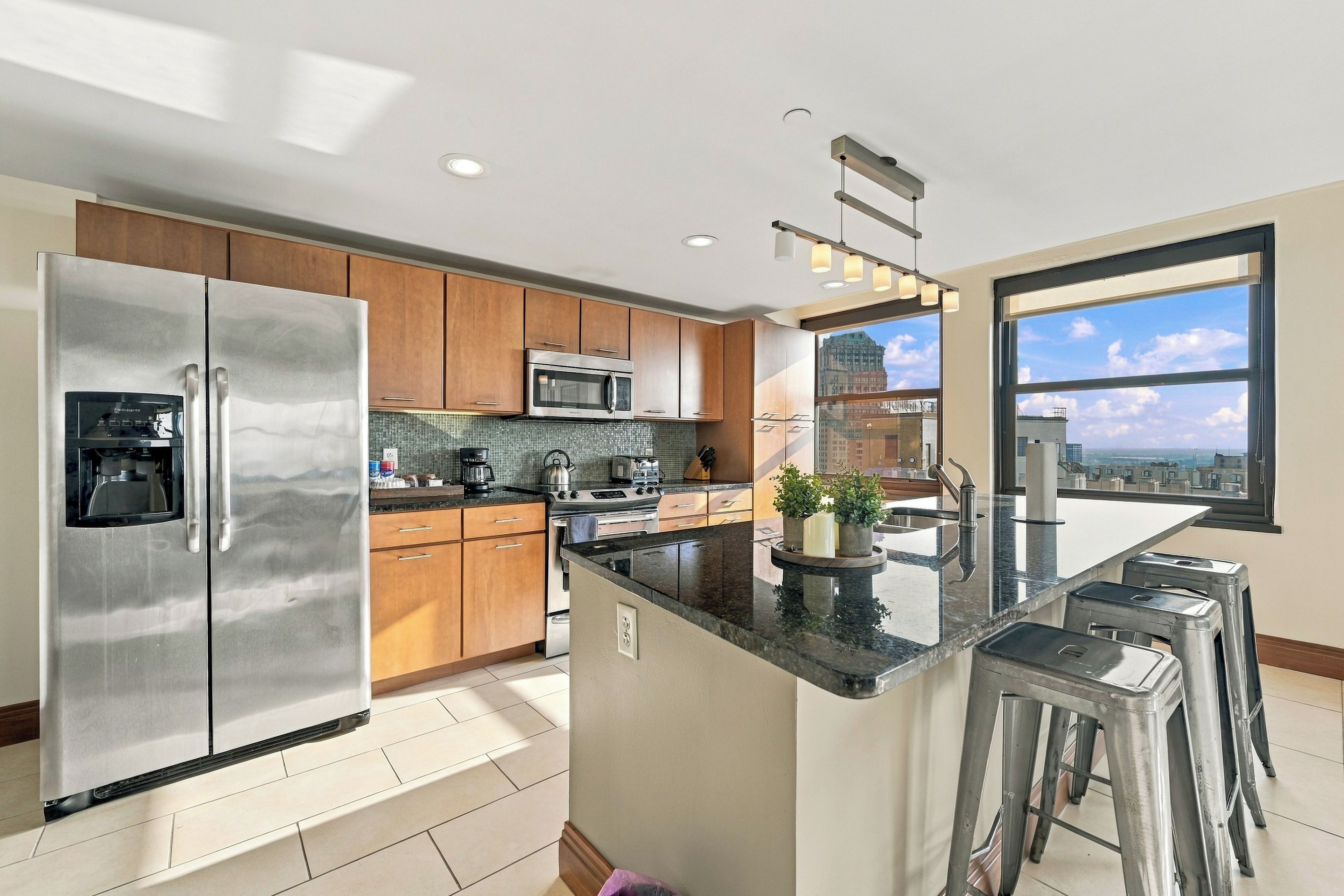 A modern kitchen with stainless steel appliances, wooden cabinets, a central island with stools, and a view of the city through large windows.