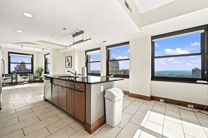 A modern kitchen with a large island, a sink, and a trash can. There are two windows providing views of the city.