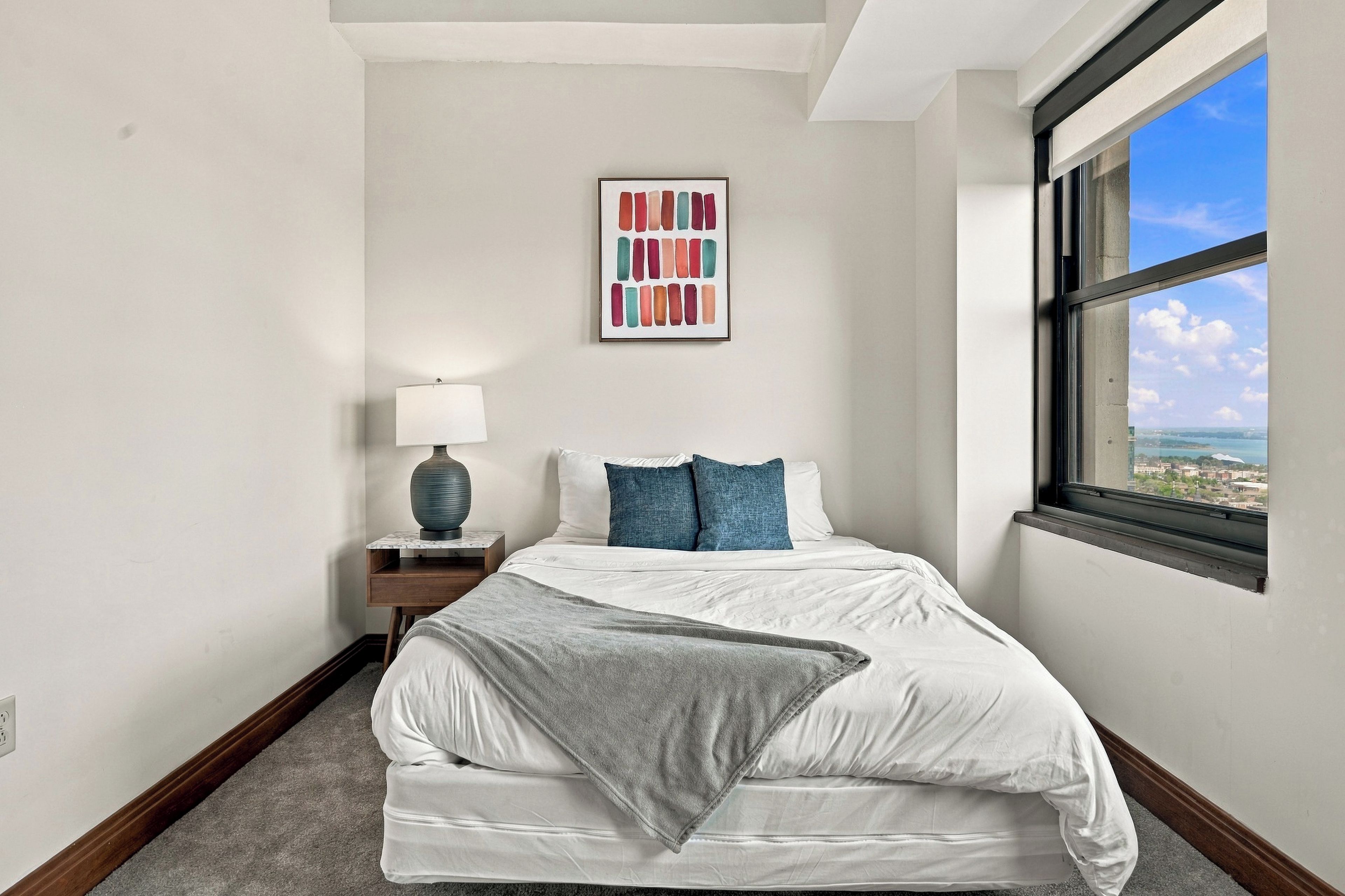Une chambre à coucher avec un lit, des oreillers, une table de chevet avec une lampe, un échantillon de couleurs encadré accroché au mur et une fenêtre donnant sur un paysage.