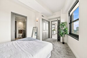 A hotel room with a bed, a bathroom, a hallway, and a window with a view of the city.