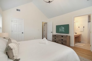 A bedroom with a large bed, a wooden dresser, a TV mounted on the wall, and a door leading to a bathroom. - Charleston Oasis With Hot Tub, Mins to Dtwn (Mount Pleasant)