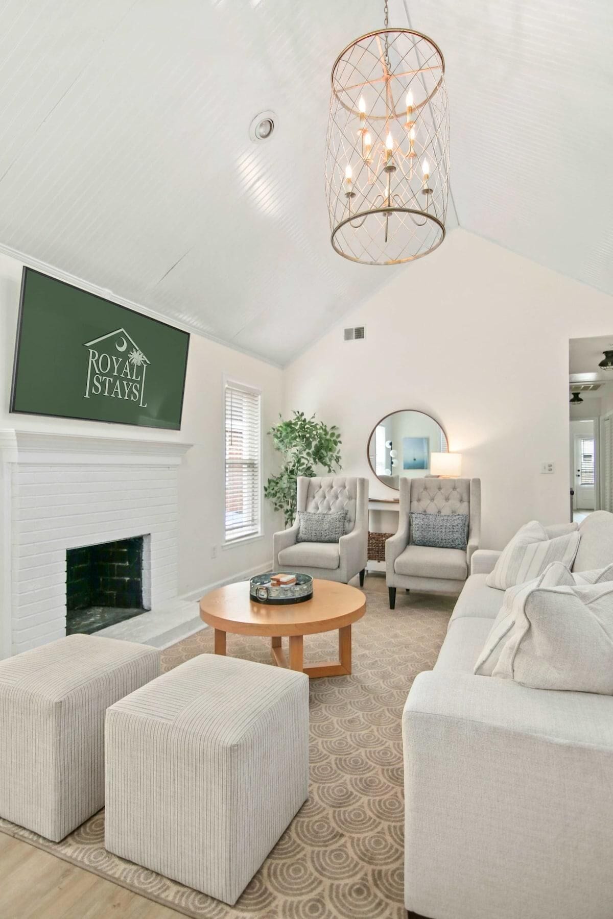 A spacious living room with a white fireplace, a large TV mounted on the wall, and a chandelier. There are two armchairs and a sofa with ottomans.