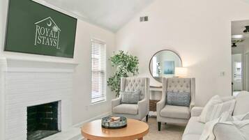 A spacious living room with a white fireplace, a large TV mounted on the wall, and a chandelier. There are two armchairs and a sofa with ottomans.