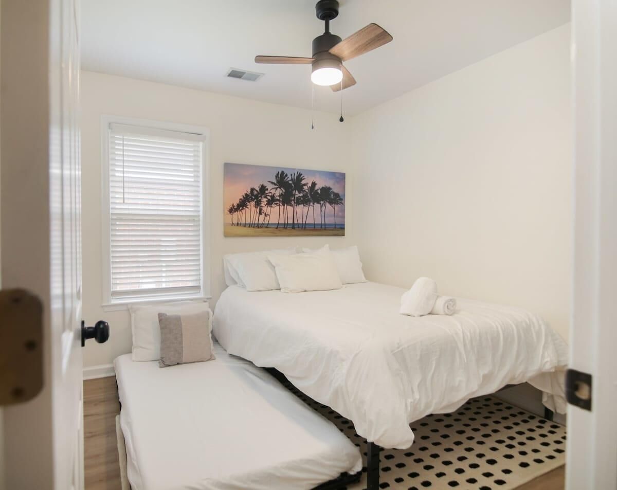 Un dormitorio con cama, un banco, un ventilador de techo y un cuadro de palmeras.