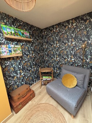 A room with a patterned wallpaper, a grey sofa with a gold cushion, a small wooden side table, and a brown suitcase. - 3-Star Vacation Rental “La maison de Doris” (Bihorel)