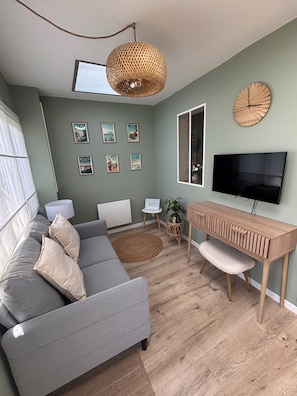 A modern living room with a grey sofa, a wooden side table, a TV, and a wall clock. - 3-Star Vacation Rental “La maison de Doris” (Bihorel)