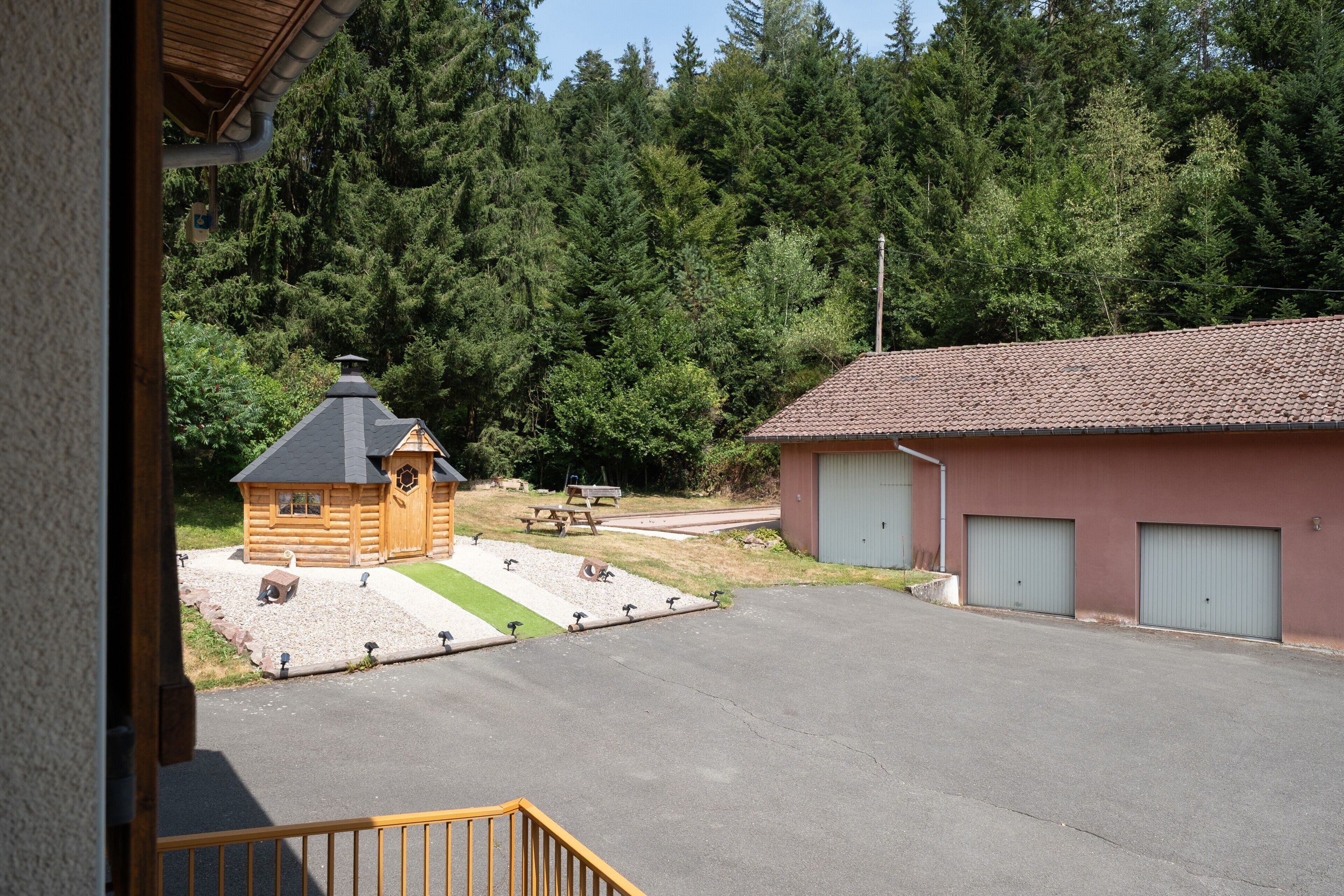 Une cabane en rondins avec un petit porche, un bâtiment rouge doté d’un garage, et une zone boisée en arrière-plan.
