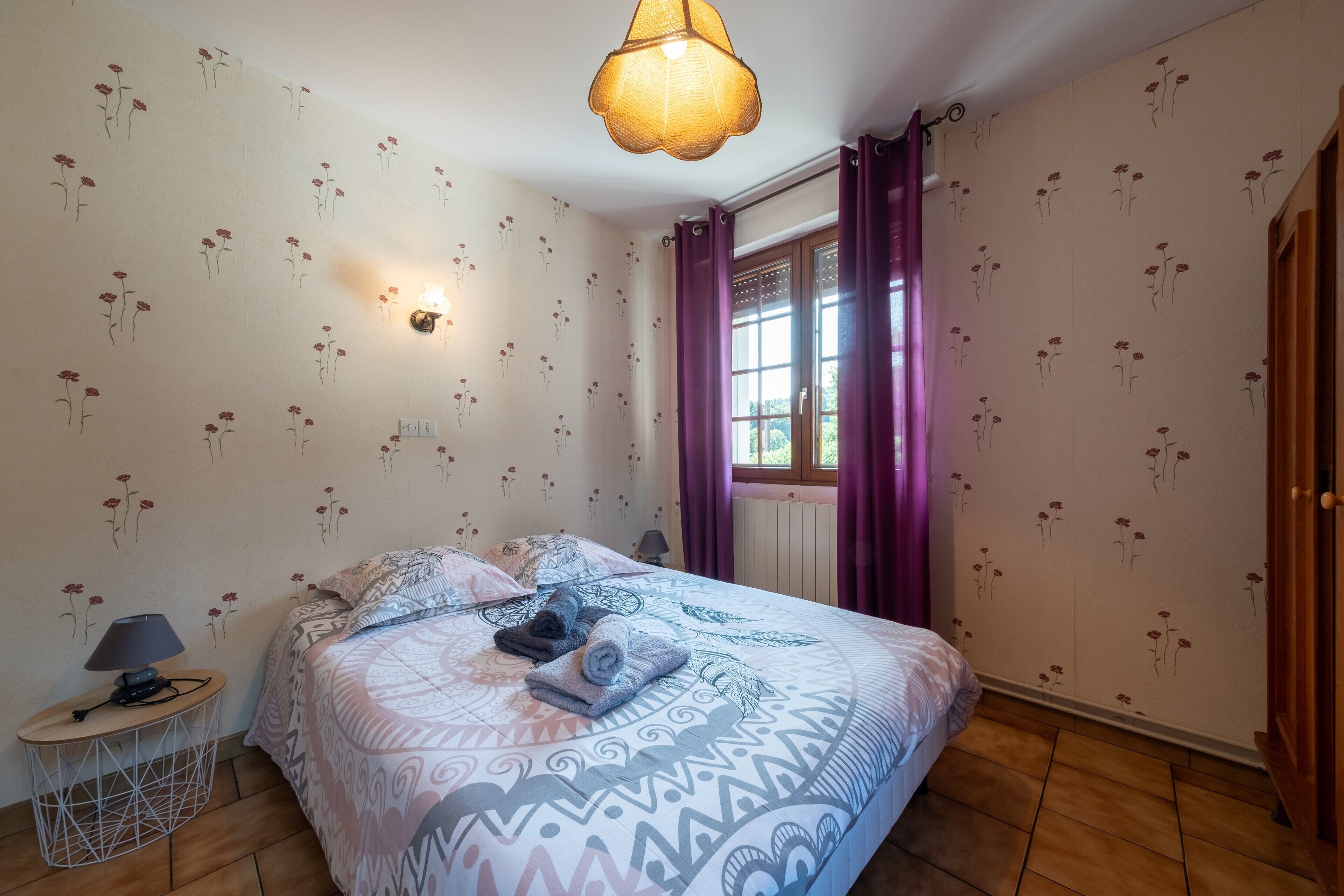 Une chambre à coucher avec une couvre-lit à motifs, une lampe de chevet et une fenêtre ornée de rideaux violets.