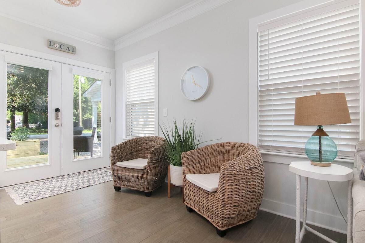 A room with two wicker chairs, a potted plant, a round wall clock, and a lamp on a small table.