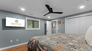 Ein Schlafzimmer mit Bett, Deckenventilator, Fenster mit Jalousien, einer Tür und einem an der Wand befestigten Schild.
