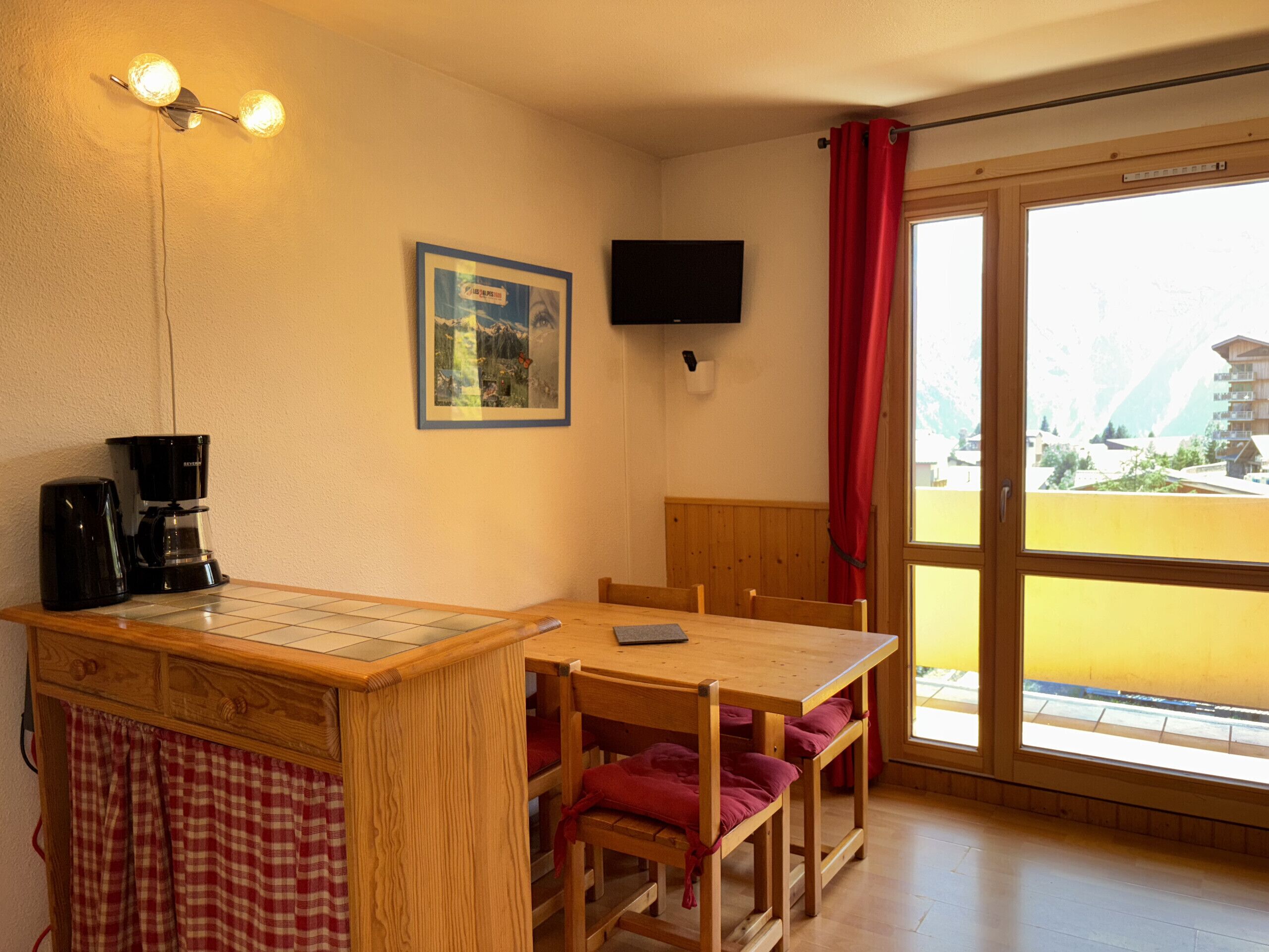 A room with a wooden table, chairs, a coffee maker, a flat-screen TV, a framed picture, and a window with a view of buildings.