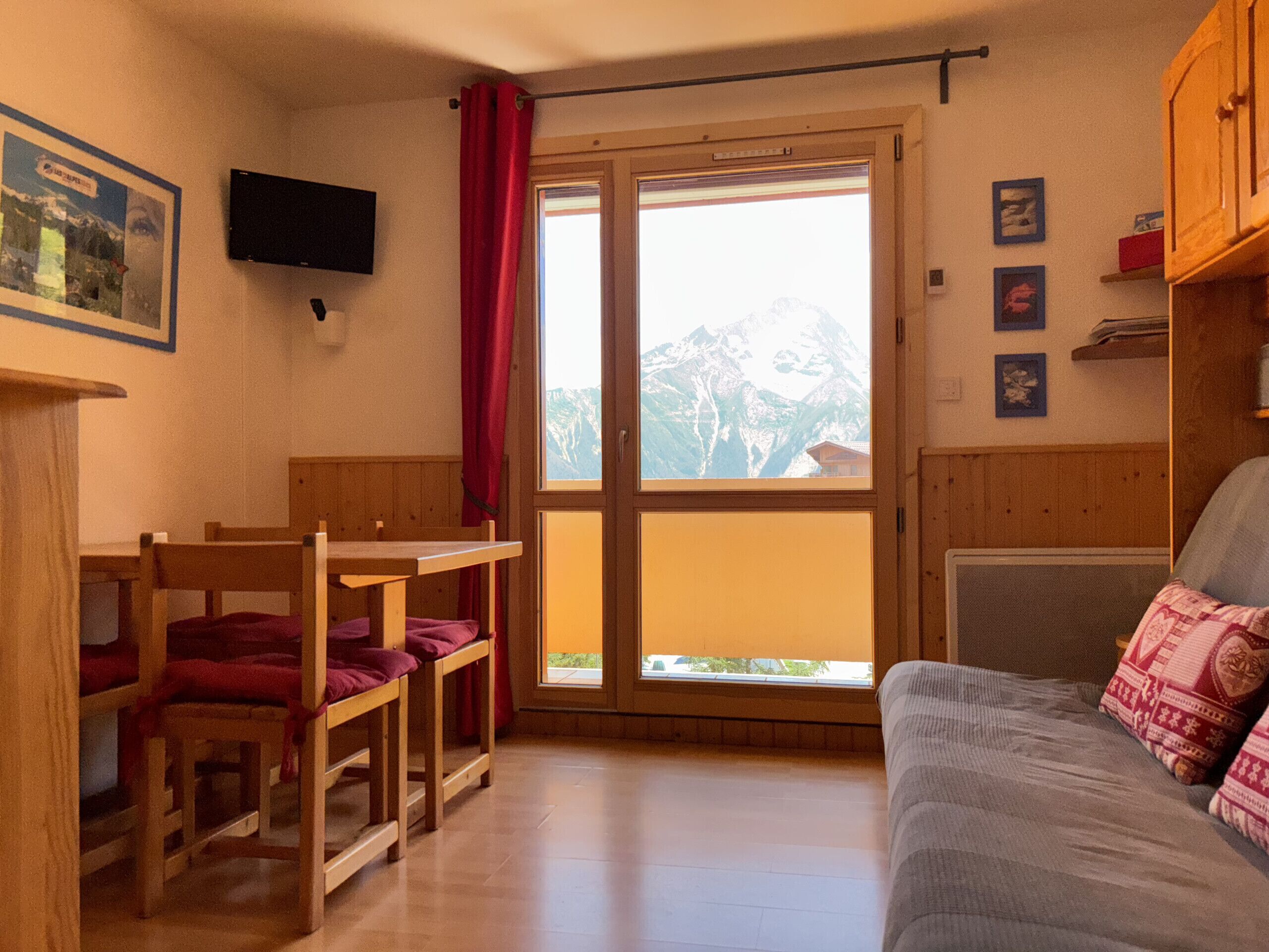 A room with a wooden table and chairs, a sofa, a TV mounted on the wall, and a window with a view of mountains.